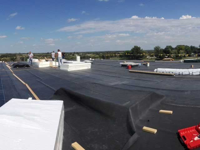 Workers on a flat roof install roofing materials under a blue sky, some white insulation is stacked.