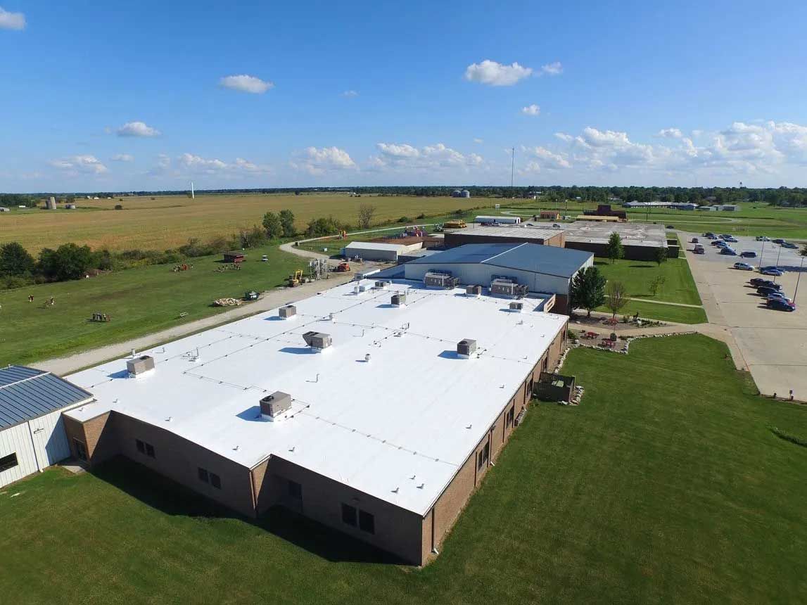 Aerial view of a long, white-roofed building on green grass, with fields and blue sky in the background.