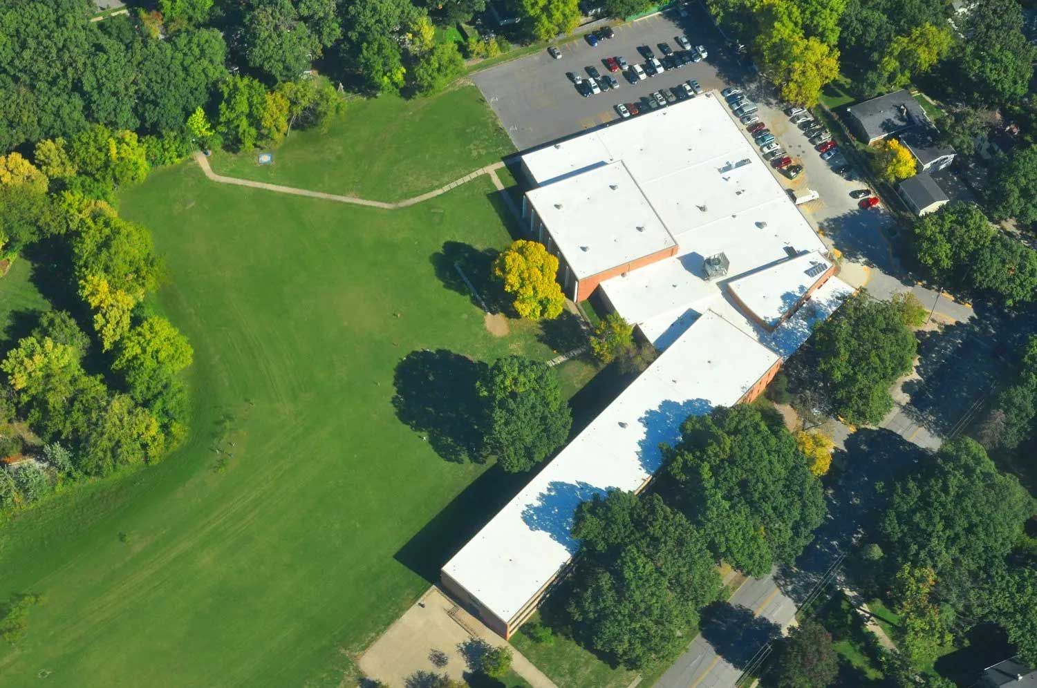 Aerial view of a school building with a white roof, surrounded by green grass and trees. A parking lot is visible.