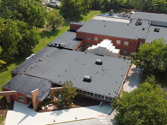 Aerial view of a school building with flat black roofs, red brick walls, and surrounding greenery.