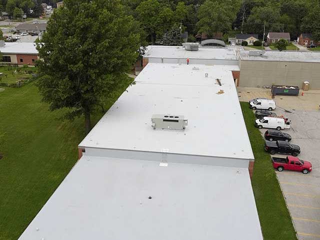 Aerial view of a flat white roof with a tree, grass, and parked vehicles.