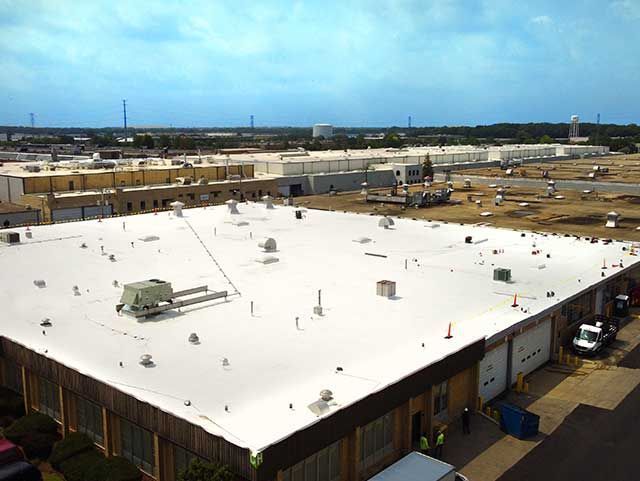 White flat commercial roof with multiple vents and equipment. Workers and a truck are at the edge. Cloudy sky.
