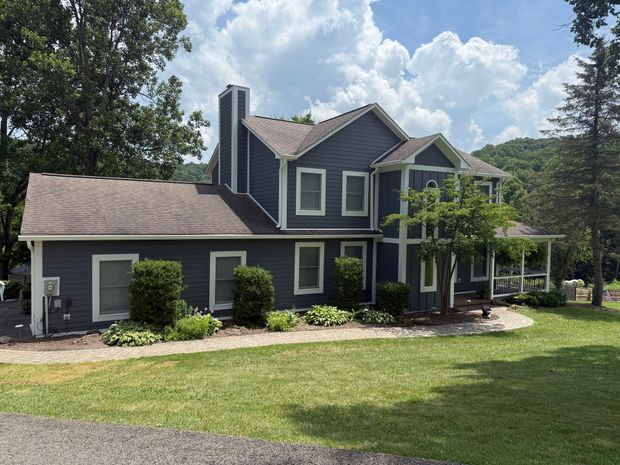 A two-story dark blue house with white trim and a stone walkway in a grassy yard under a bright, cloudy sky.