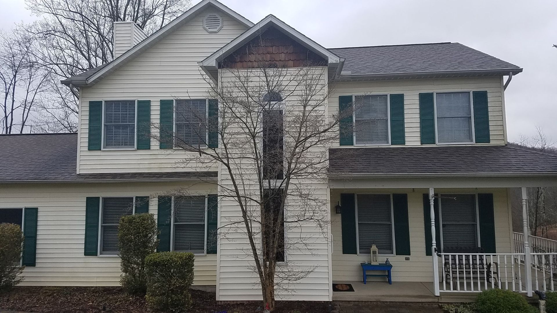 A two-story house with beige siding, dark green shutters, and a central protruding entryway under a cloudy sky.