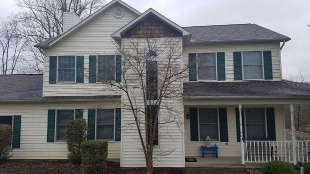 A two-story house with beige siding, dark green shutters, and a central protruding entryway under an overcast sky.