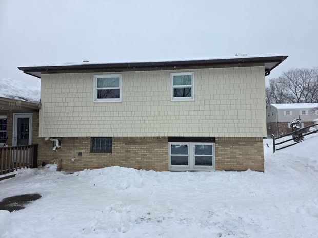 A side view of a house with light-colored square shingles over a brick foundation in a snowy, outdoor setting.