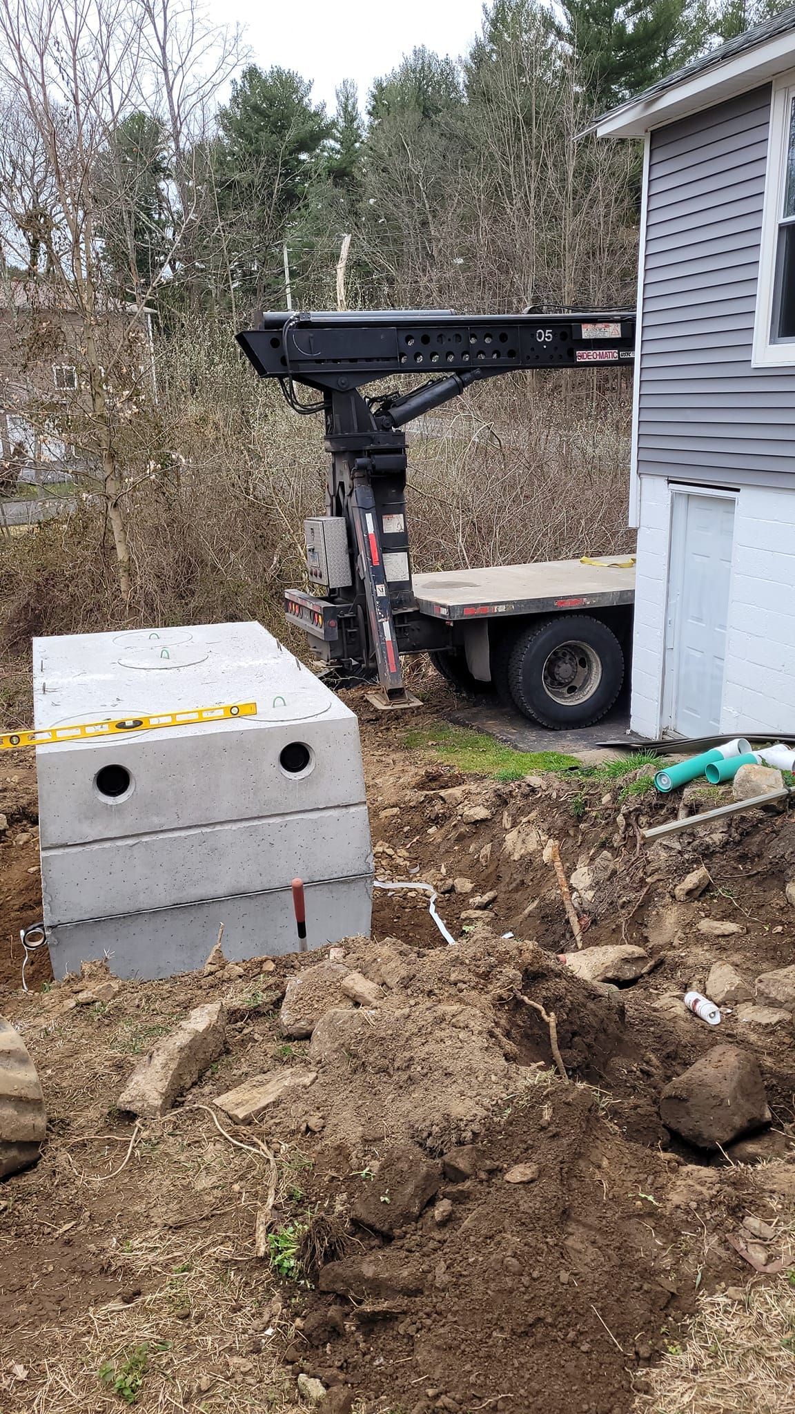 A crane is lifting a concrete block in the dirt in front of a house.