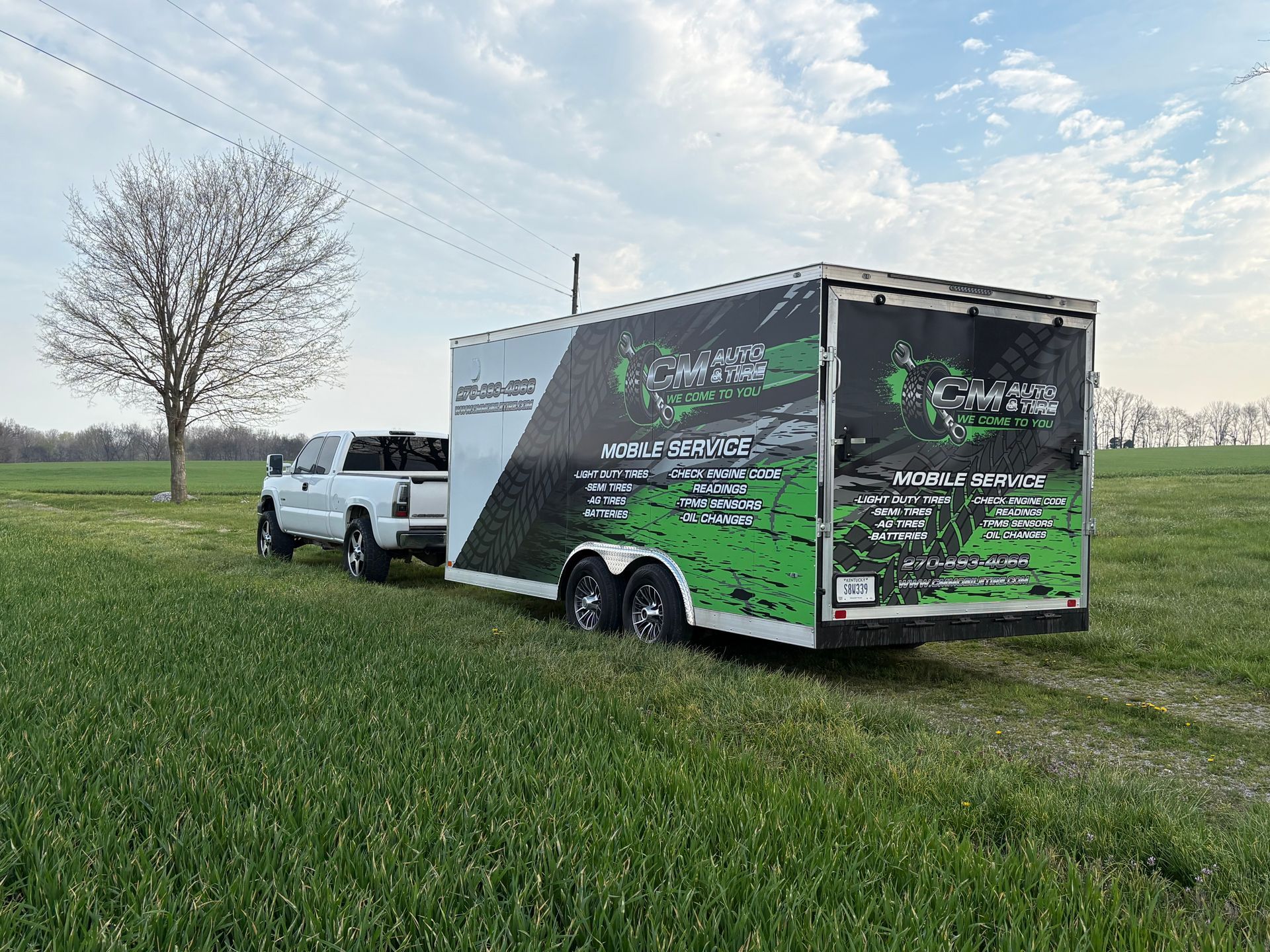 White truck towing a black-and-green enclosed trailer on a grassy field under a cloudy sky