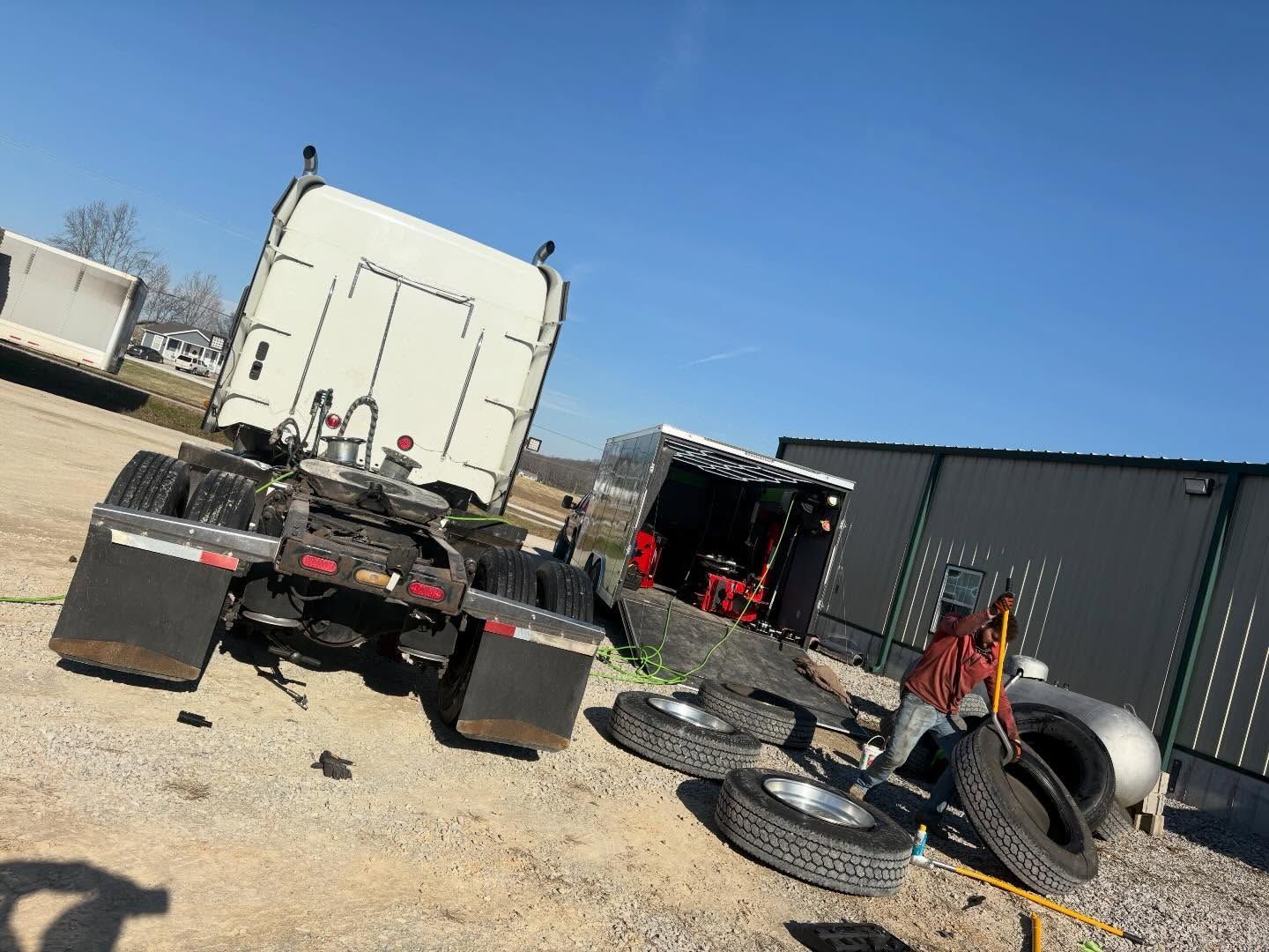 Truck chassis and loose tires in a gravel yard beside a white trailer and metal building under a blue sky