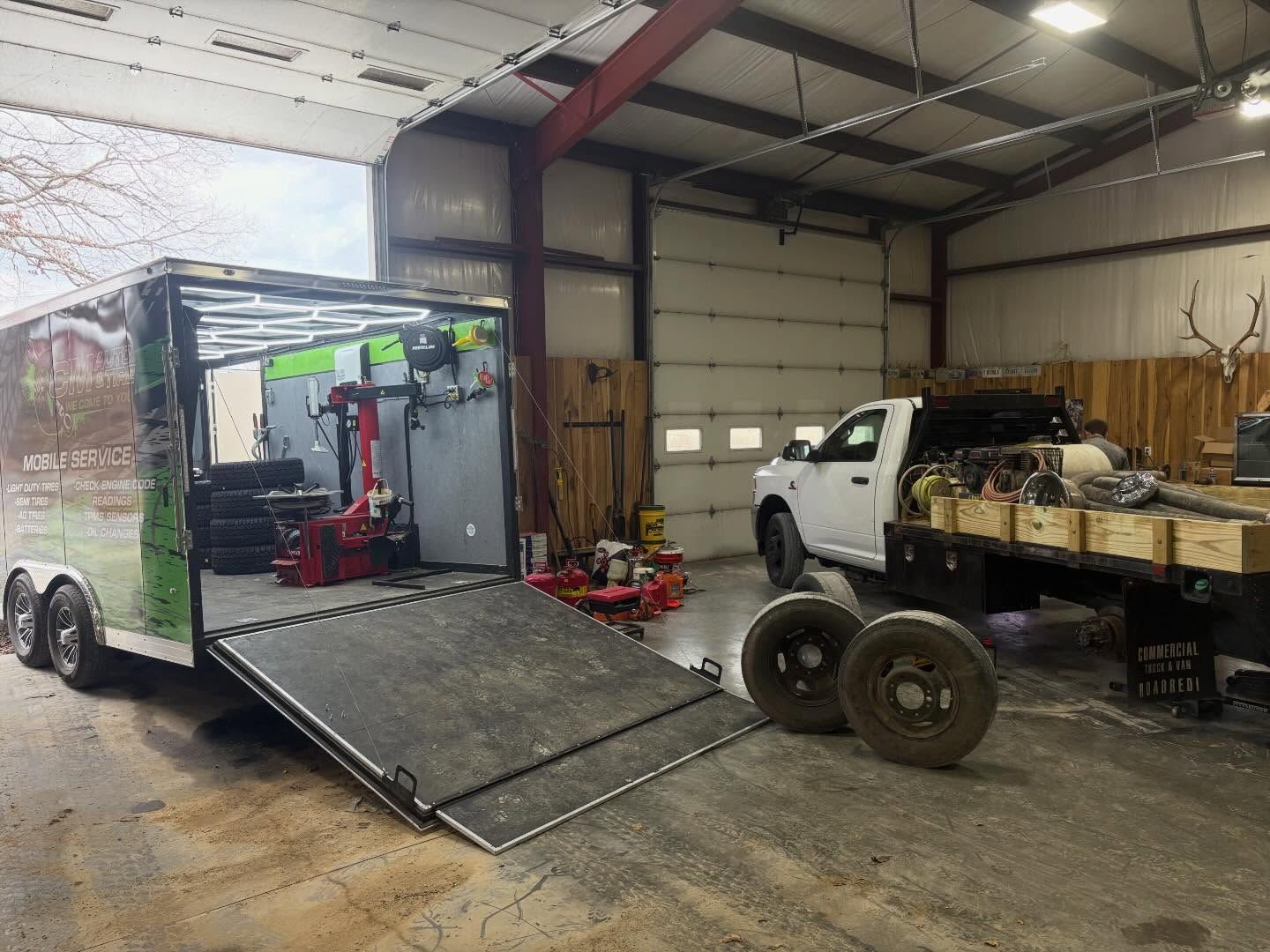 White pickup truck and enclosed trailer with ramp in a workshop, alongside loose wheels and tools.