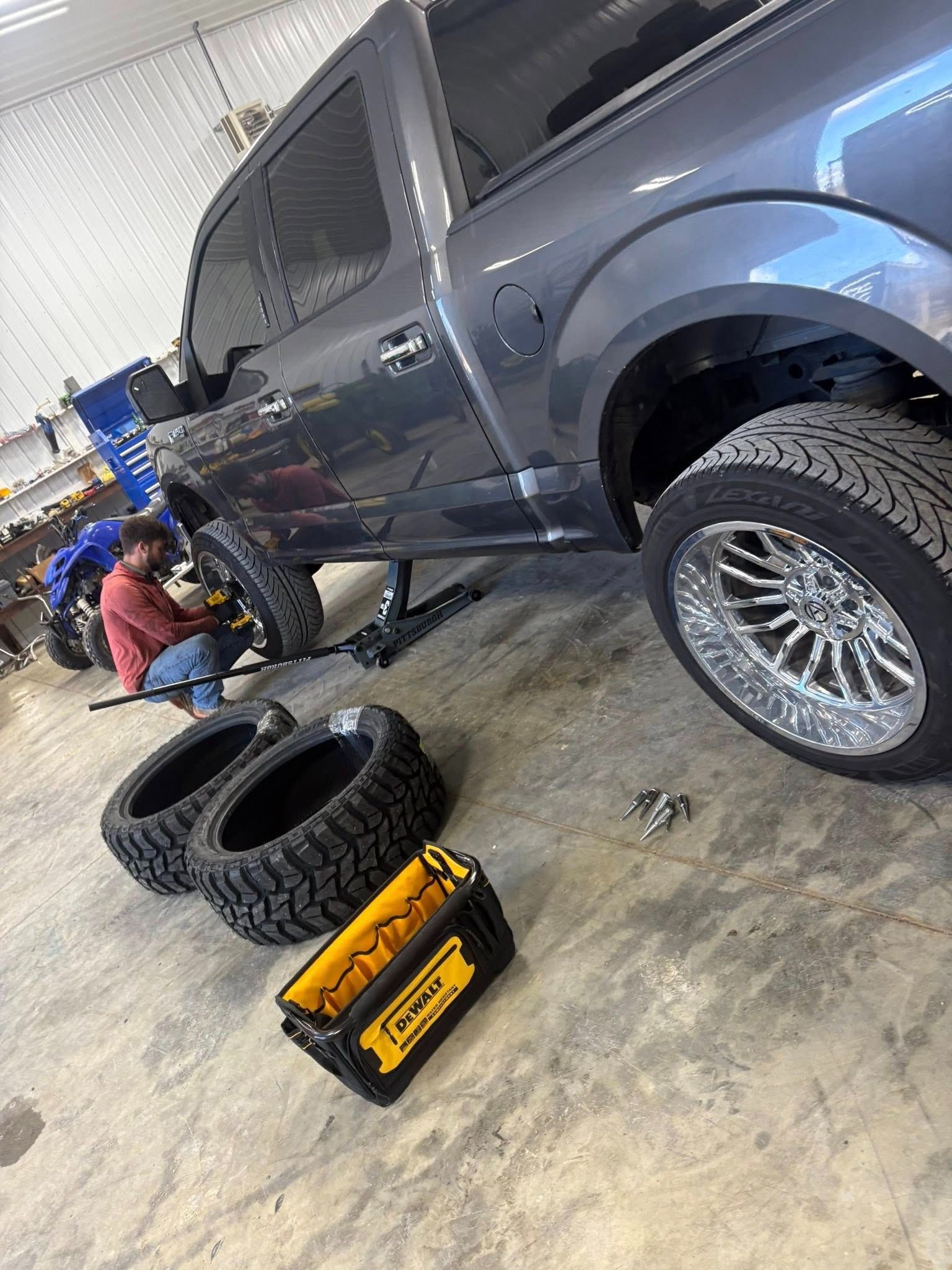 Mechanic working on a gray pickup truck in a garage, with tires and a tool case on the floor.