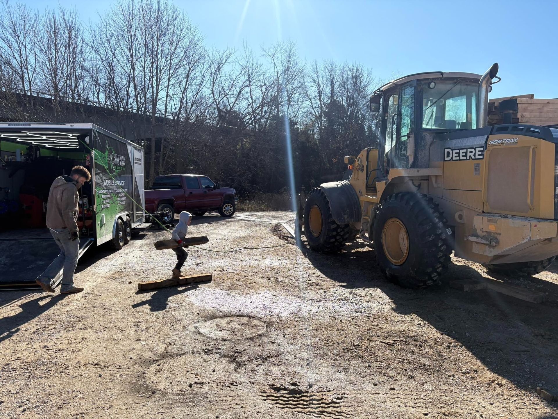Backhoe parked on muddy lot near shed, with a person and dog standing nearby in winter sunlight