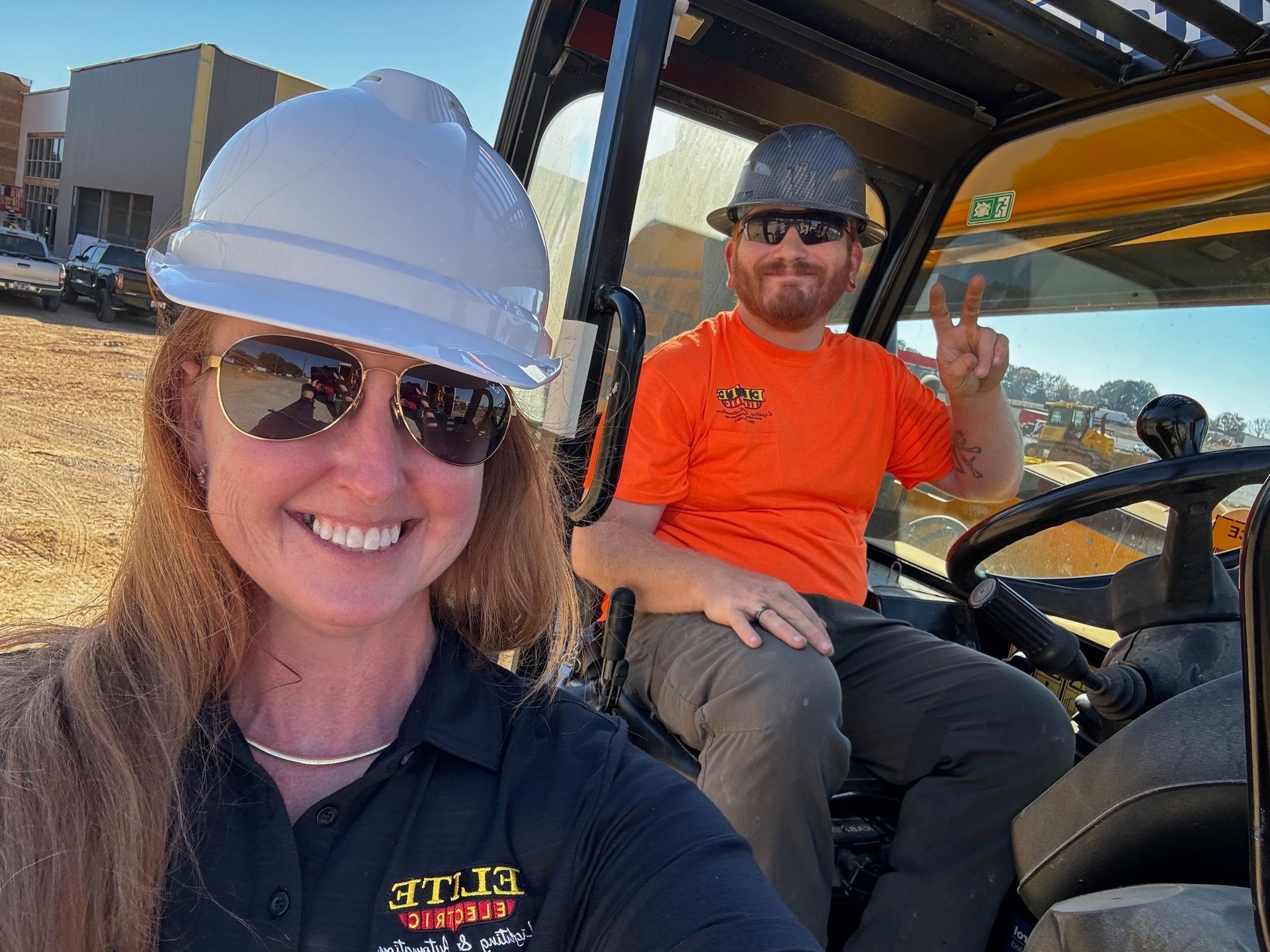 A man and a woman are sitting in a forklift . the woman is wearing a hard hat and sunglasses.