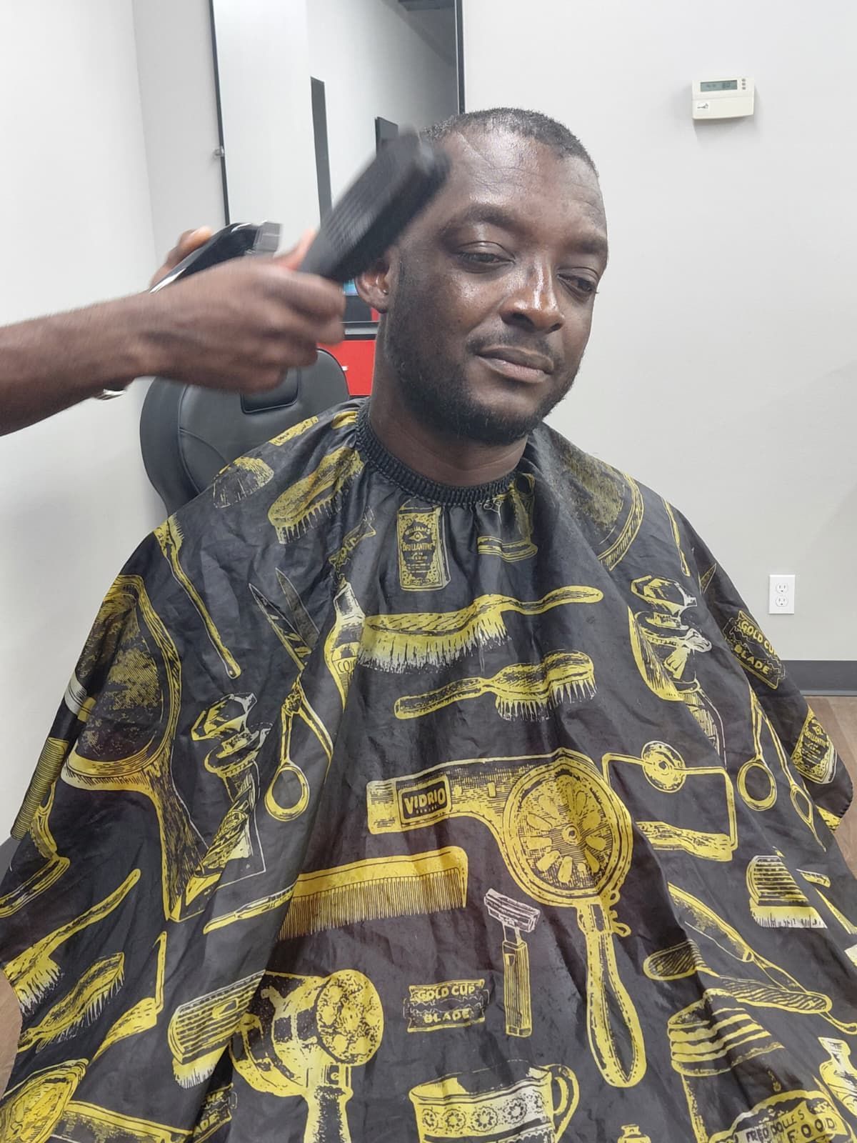 Man getting a haircut, seated and wearing a cape in a barbershop.