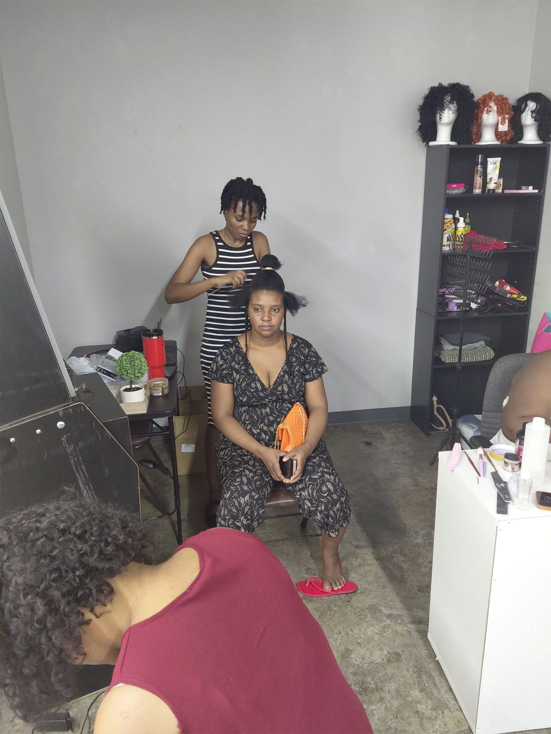 Three women, one styling hair of another seated, in a room with supplies and wigs.
