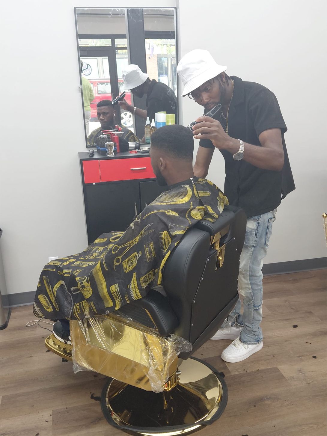 Barber cutting a customer's hair in a shop. Both men are Black. The customer is seated in a gold-accented chair.
