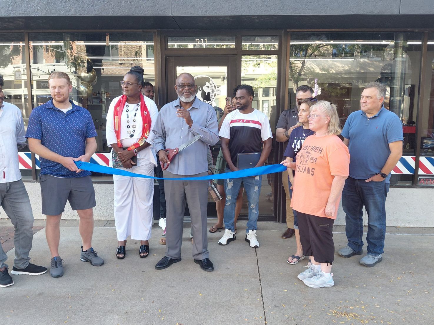 Group cuts blue ribbon at a barbershop opening; diverse people, sidewalk setting.