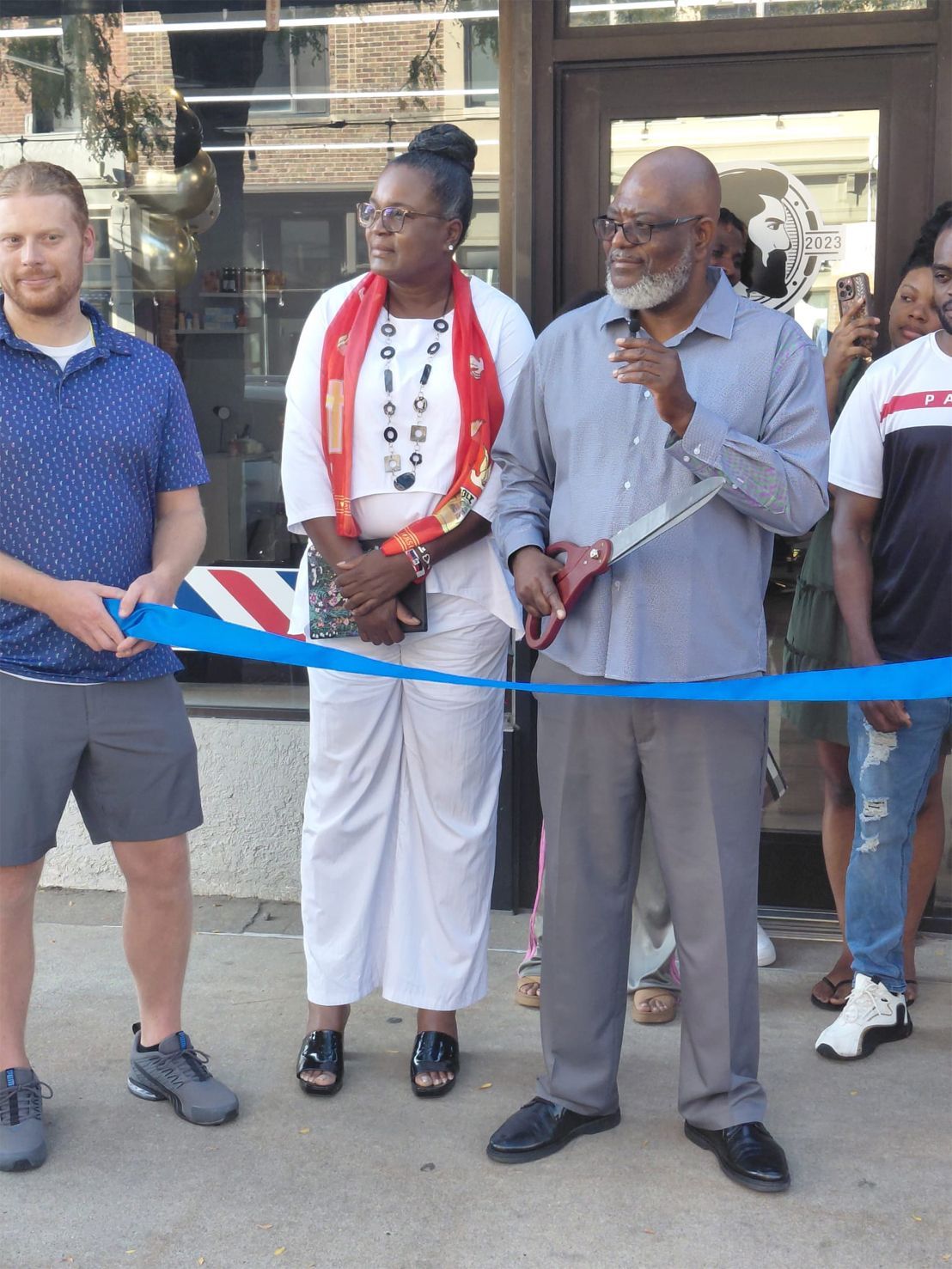 People cutting a blue ribbon at a store opening. Man speaks, woman watches, others stand.
