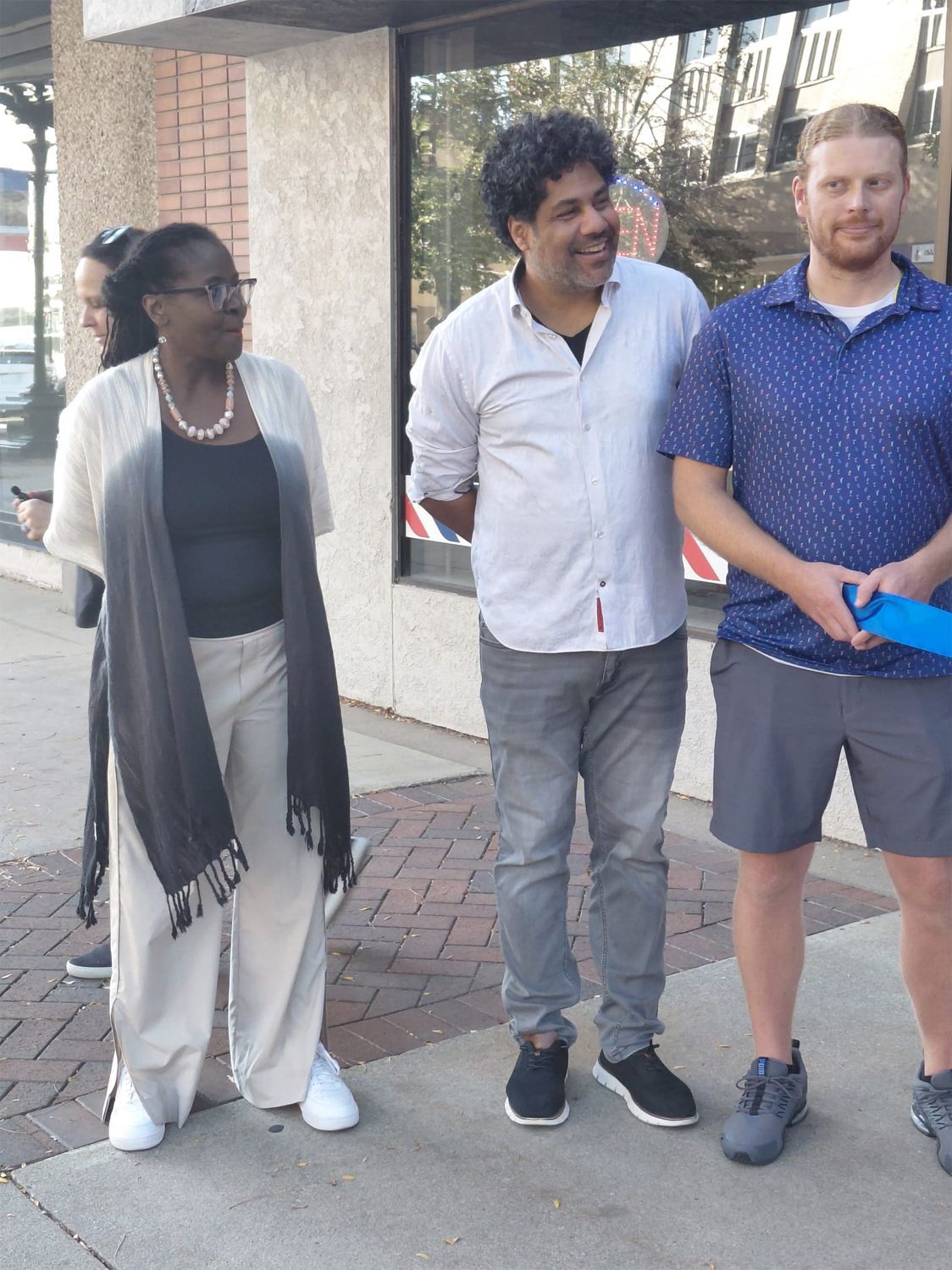 Group of four people standing on a sidewalk. Woman in white pants and black/white shawl; two men smiling, one holding blue item.