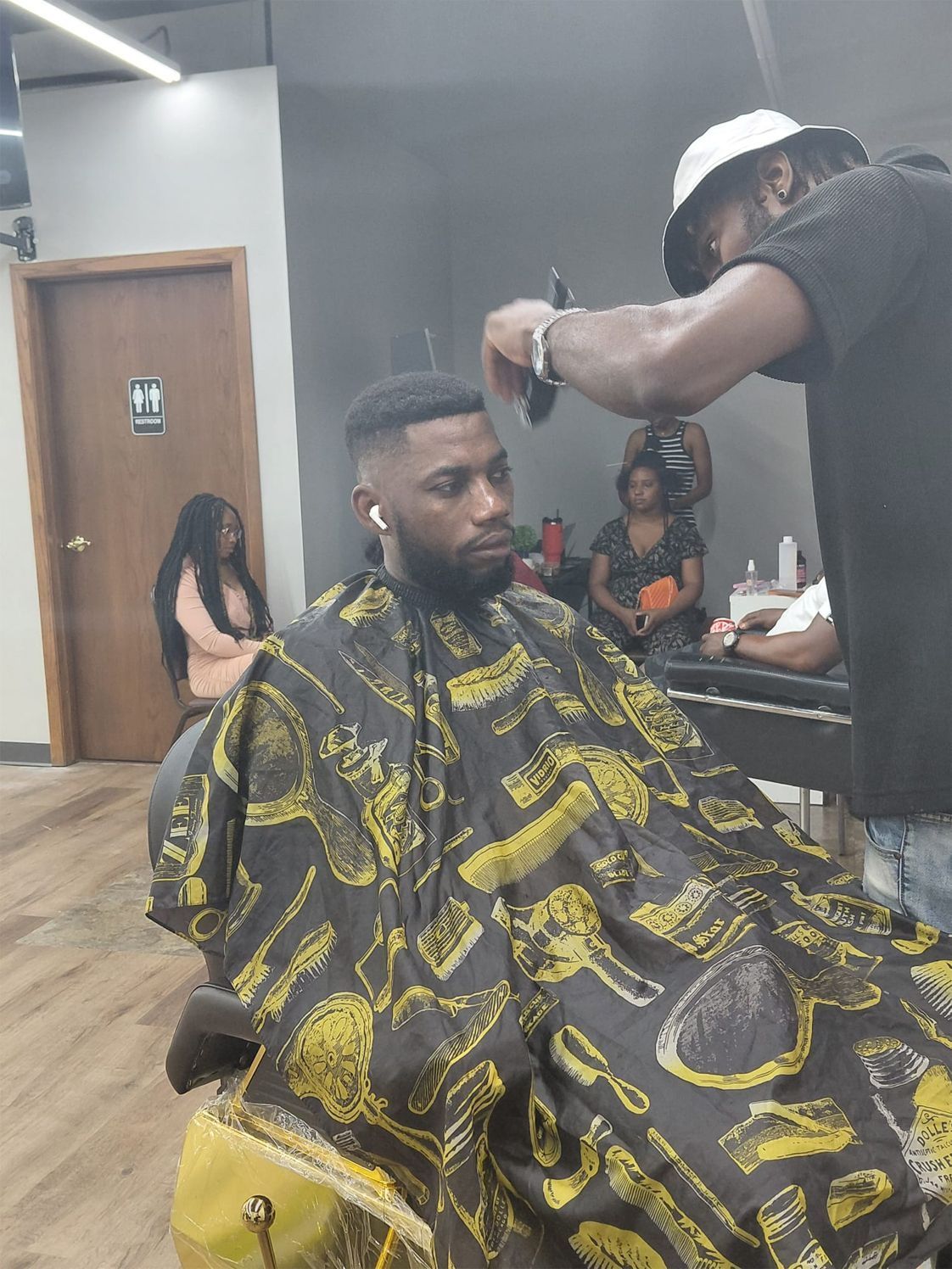 Man getting a haircut at a barbershop. Barber with hat, using clippers. Others seated in background.
