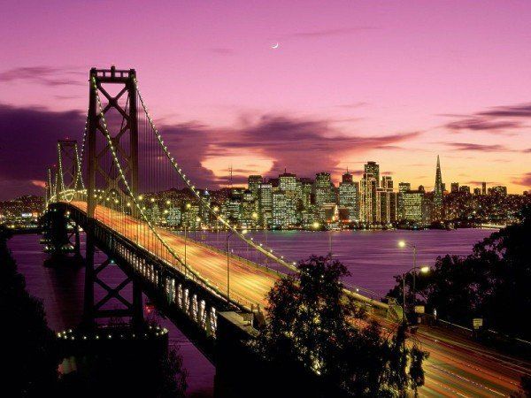 Suspension bridge lit at dusk with city skyline and purple sky in the background
