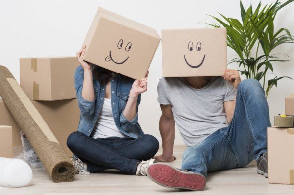 Two people sitting among moving boxes, wearing cardboard box masks with smiley faces in a room