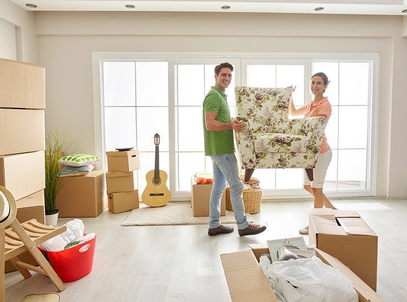 Couple carrying a floral armchair in a bright room with moving boxes and packed belongings