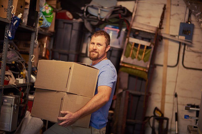 Man carrying stacked cardboard boxes in a cluttered workshop or storage room