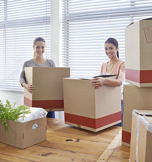 Two people carrying moving boxes in a bright room with blinds and stacked packages.