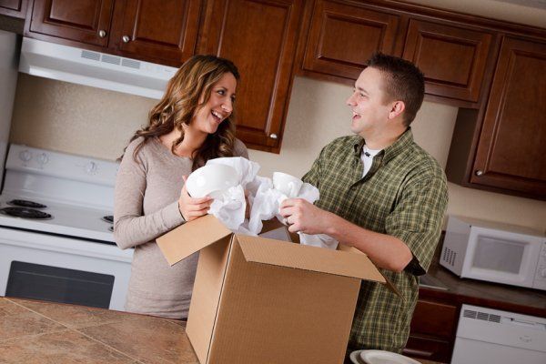 Two people unpacking dishes from a box in a kitchen, smiling at each other.