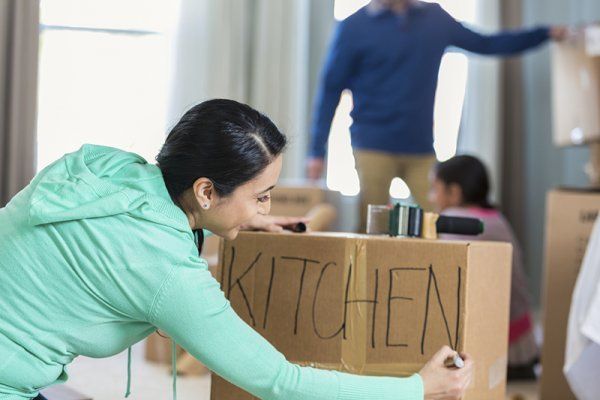 Woman in a mint jacket labels a cardboard box “KITCHEN” in a moving room.
