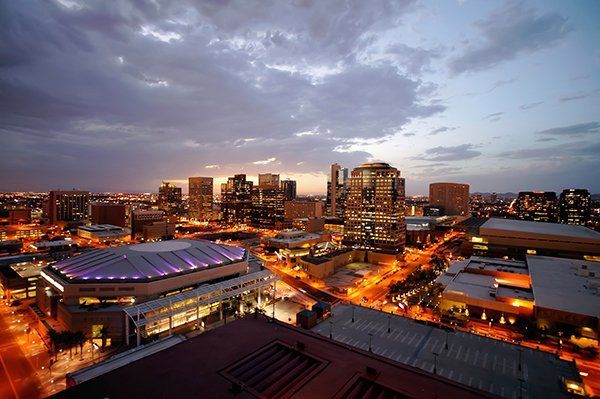 City skyline at dusk with lit buildings and a purple-roofed arena under a cloudy sky