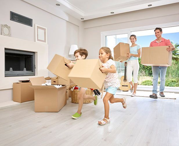 Family carrying cardboard boxes through a bright empty living room during a move