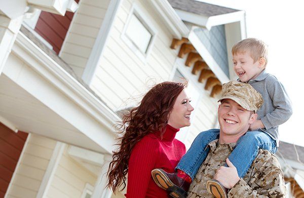 Family in winter clothing smiling outside a house, with a child on a man's shoulders.