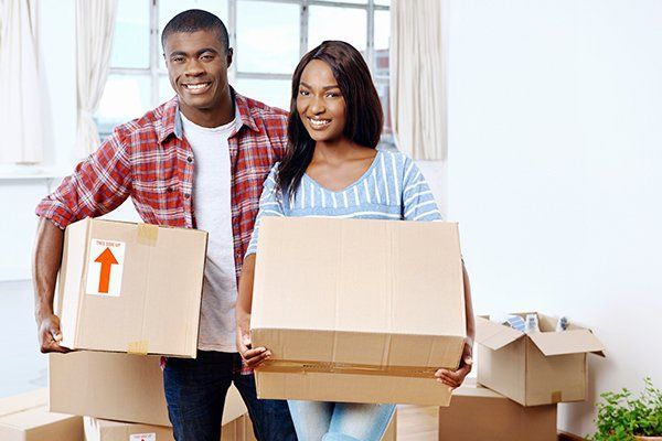 Two people carrying moving boxes in a bright room, smiling at the camera.