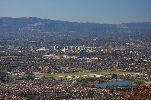 Wide cityscape with dense buildings, a runway, and a blue lake beneath distant mountains