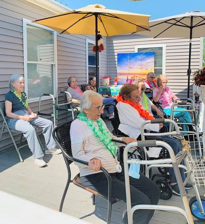 Group of seniors seated outdoors in folding chairs under umbrellas, socializing on a sunny patio.