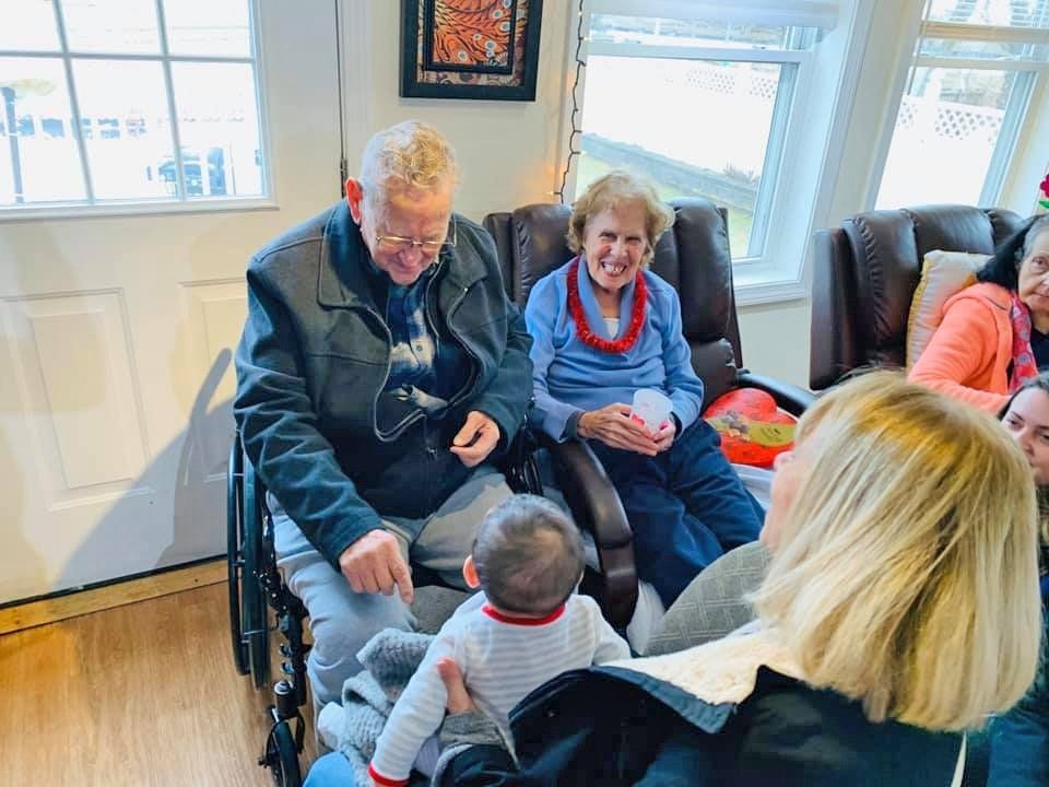 Elderly couple seated in a living room, smiling at a toddler facing them.