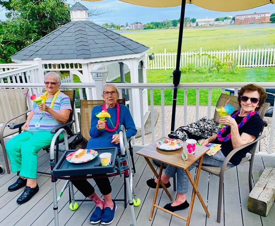 Three people sit on a deck eating colorful drinks and snacks, with a white fence and grassy field behind them.