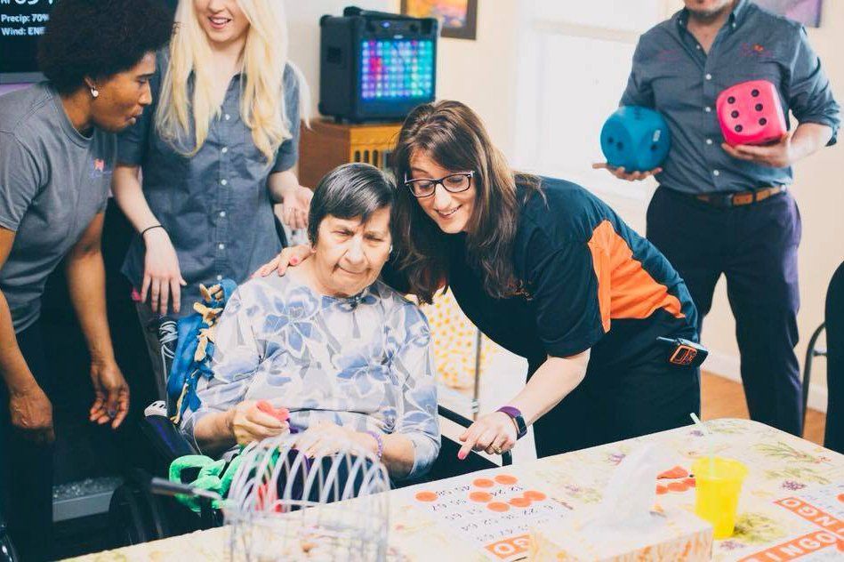 People gathered around a table, watching an older woman arrange colorful craft materials