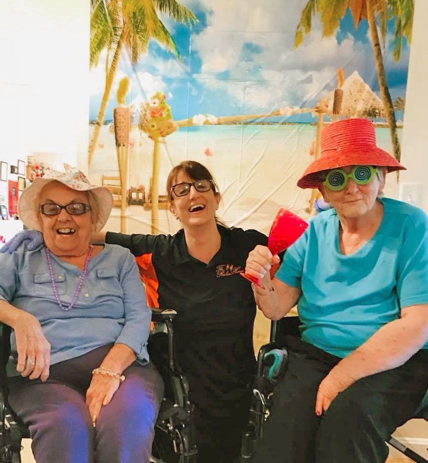 Three smiling people in hats sit in wheelchairs before a tropical beach mural