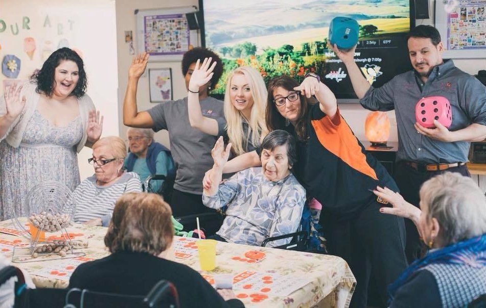 People celebrating at a table, smiling and holding up colorful painted hands and a pink die in a bright room