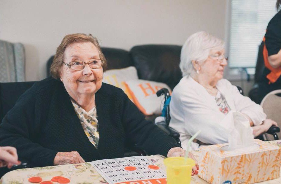 Two women seated at a table in a community room, smiling, with tablecloth, cups, and gift bags.