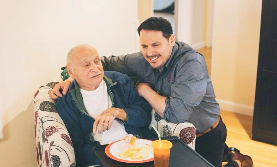 Two people smiling beside a seated elderly man with a meal and drink in a living room