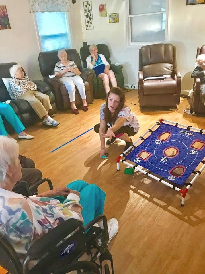 People seated in a living room watch a woman lean over a blue tabletop game.