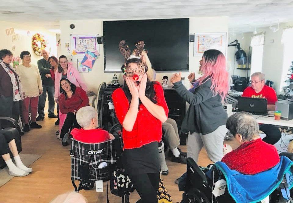 Performer in red shirt singing to a small audience in a bright room, with seated and standing listeners.