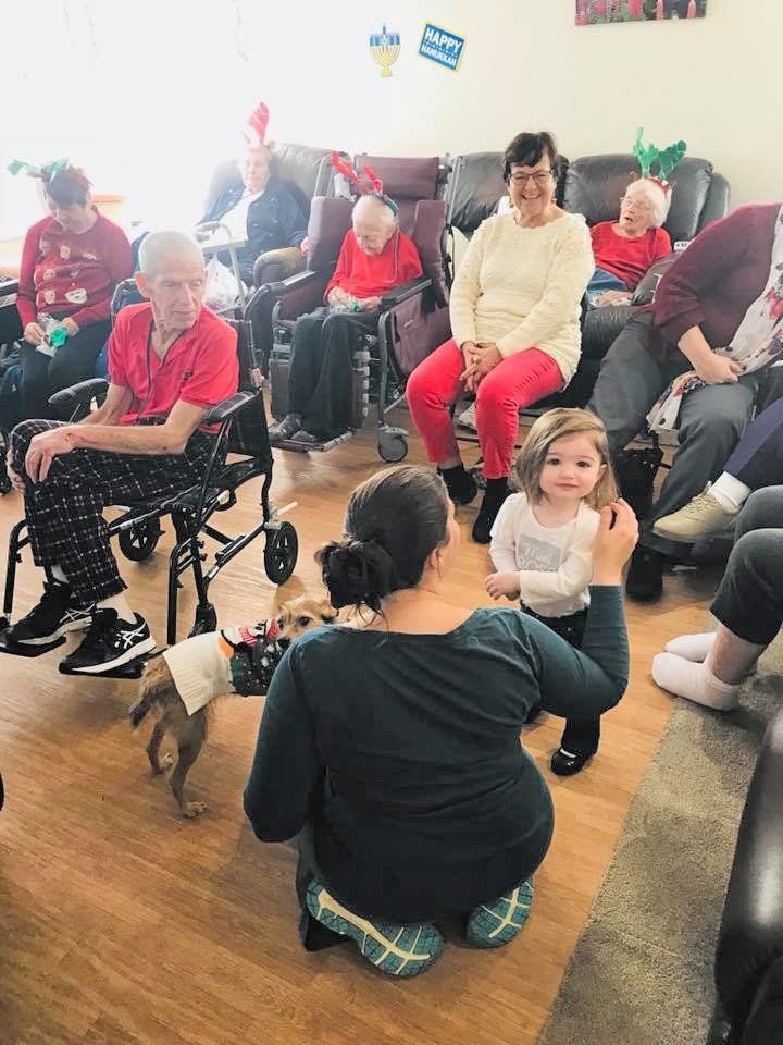 Group seated in a cozy room, with a child playing on the floor among adults and seniors.