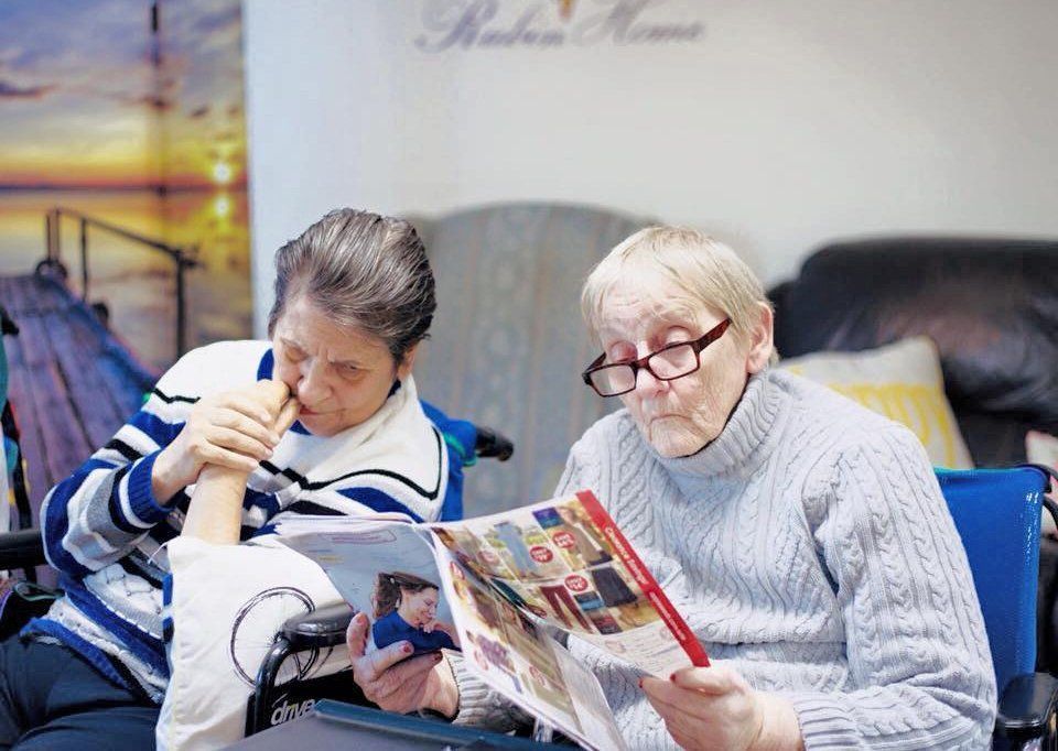 Two women reading magazines on a couch, one wearing glasses in a light sweater.