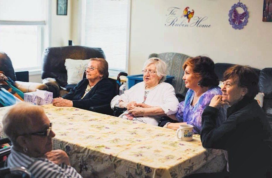 Several people sit around a table in a bright living room, chatting during a gathering.