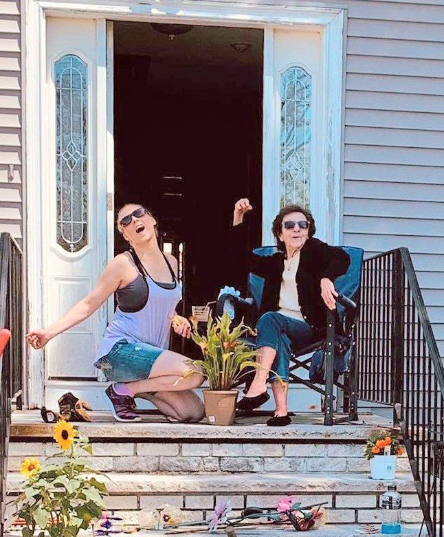 Two people posing on a front porch step, one kneeling and one seated, with potted plants and flowers nearby.